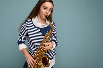 Shot of pretty positive happy brunet little girl wearing stylish striped longsleeve standing isolated over blue background wall playing saxophone looking at camera