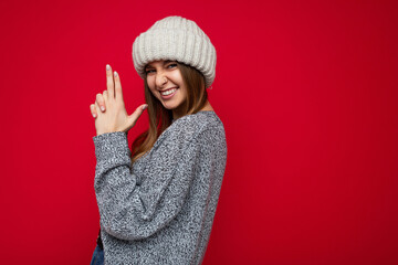Portrait of positive happy young beautiful dark blonde woman with sincere emotions wearing grey sweater and beige knitted hat isolated over red background with copy space and making shooting gun with
