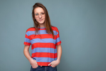 Photo shot of beautiful young brunette woman wearing optical glasses casual srtiped pink and blue t-shirt standing isolated over blue background wall and looking at camera