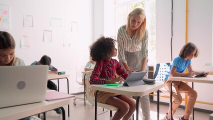 Happy smiling Caucasian teacher explaining encouraging Afro American schoolgirl using tablet device sitting in classroom with group of diverse schoolchildren. Modern education technologies concept. - Powered by Adobe