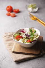 A small white salad bowl with microgreens, cherry tomatoes and fresh basil leaves on a wooden board on a grey concrete surface