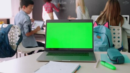 Laptop computer green blank empty mockup screen on school desk with elementary junior children students in classroom background. Education software website technology ads concept.