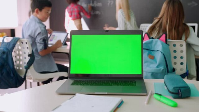 Laptop computer green blank empty mockup screen on school desk with elementary junior children students in classroom background. Education software website technology ads concept.