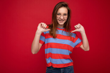 Photo of positive happy young beautiful brunette woman with sincere emotions wearing casual striped t-shirt and optical glasses isolated over red background with empty space
