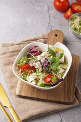 A small white salad bowl with microgreens, cherry tomatoes and fresh basil leaves on a wooden board on a grey concrete surface