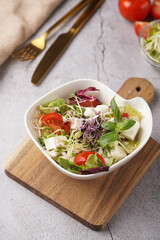 A small white salad bowl with microgreens, cherry tomatoes and fresh basil leaves on a wooden board on a grey concrete surface