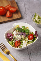A small white salad bowl with microgreens, cherry tomatoes and fresh basil leaves on a wooden board on a grey concrete surface