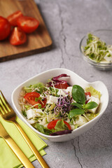 A small white salad bowl with microgreens, cherry tomatoes and fresh basil leaves on a wooden board on a grey concrete surface