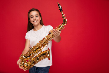 Fototapeta na wymiar Photo shot of beautiful happy smiling brunette little girl wearing white t-shirt for mockup standing isolated over red background wall holding musical instrument naming saxophone looking at camera