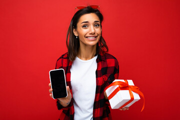 Beautiful happy young brunette woman isolated over red background wall wearing white casual t-shirt and red and black shirt holding white gift box with red ribbon and mobile phone with empty screen