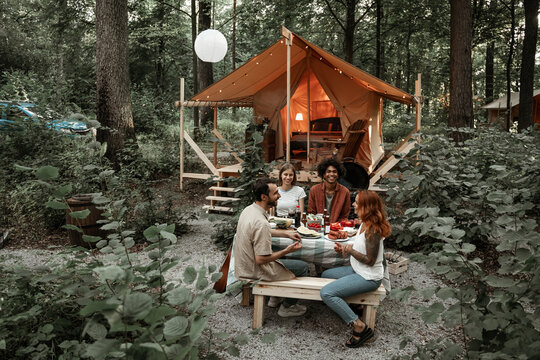 Young friends having dinner at glamping talking, laughing after sunset. Happy millennial gang camping at open air picnic under bulb lights. Spending time with friends outdoors, barbeque party
