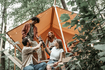 Young friends enjoy sunny evening in forest glamping. People laugh and toast with beer cider bottles and wine glasses by the bonfire near tent. Camping hanging out together, social gathering outdoors