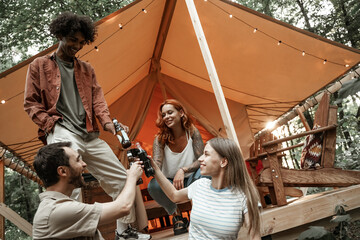 Young friends enjoy sunny evening in forest glamping. People laugh toast clinking beer cider bottles and wine glasses by bonfire near tent. Camping hanging out together, social gathering outdoors