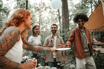 Group of happy young friends making barbecue in forest in glamping. African man grilling sausages passing camp food to his friend, laughing, having fun on a picnic in the countryside, grilling