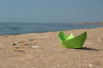 Fototapeta na wymiar Light green paper boat on sandy beach near sea, space for text