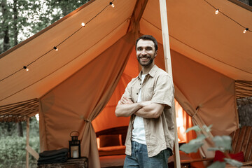 Young smiling man standing inside glamping tent looking out at forest nature. Outdoors wooden cabin with light bulbs. Cozy, camping, glamping, holiday, vacation lifestyle concept.
