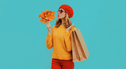 Autumn portrait of beautiful young woman with shopping bags and yellow maple leaves wearing a knitted sweater and red beret on blue background