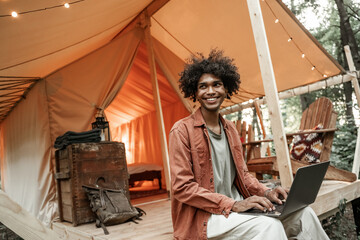 Young smiling African man with piercing sitting at glamping typing on laptop. Camping lifestyle. Low budget travel, holiday. Wi-fi connection information communication technology. Remote work - obrazy, fototapety, plakaty