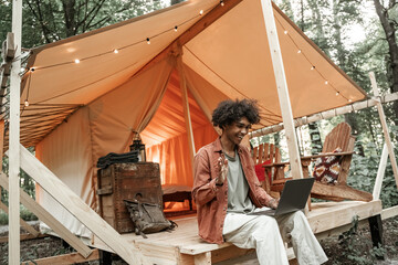 Young smiling African man with piercing talking on video-call at a laptop. Camping lifestyle. Low budget travel, holiday in forest. Wi-fi connection information communication technology. Remote work
