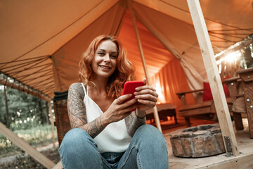Red-haired smiling girl sits next to the tent and chatting messaging streaming on the smartphone, posting, liking in social media. Travel outside city in the woods. Camping.