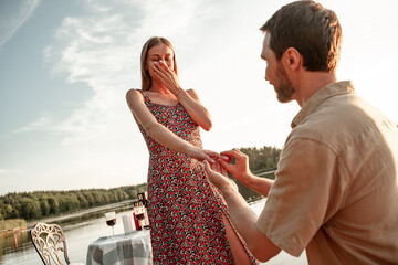 Handsome young man putting engagement ring on hand of beloved pretty girlfriend, asking to marry. Guy standing on his knee during sunset offering engagement ring to excited woman on sky background - obrazy, fototapety, plakaty