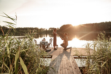Handsome young man requesting hand of beloved pretty girlfriend, asking to marry. Guy standing on his knee during sunset offering engagement ring to surprised woman outside on the forest lake - obrazy, fototapety, plakaty