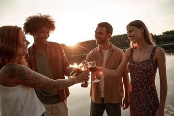 Group of friends having fun on picnic near a lake or river, drinking wine, beer, cider clinking glasses while saying a toast. Smiling young people partying outdoors during sunset in countryside.