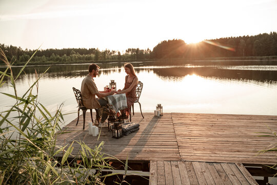 Young couple spend time together drinking wine on wooden pier: handsome man holds his girlfriend hand, making marriage proposal during sunset, wearing engagement ring, forest lake background