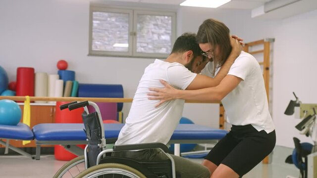 Handheld shot of a Caucasian male disabled patient, and female physiotherapist, exercising a proper lifting technique from a wheelchair, with squatting down, bending at the hips and knees only