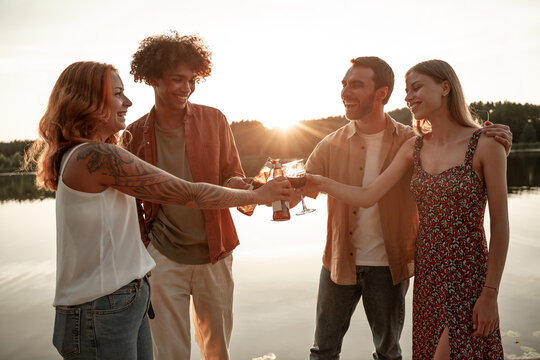 Young adults multiracial friends couples having fun, celebrating together clinking red wine, beer, cider outdoors on pier at sunset background drinking together, laughing, cheers. After lockdown