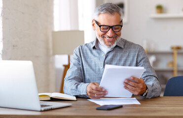 Handsome smiling senior man reading financial documents while working remotely on laptop at home