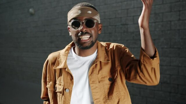 Talented African American man wearing trendy clothing and sunglasses is singing and gesturing looking at camera against urban background on sunny day.