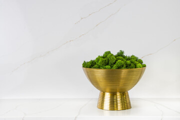 A bunch of green moss being placed in a golden brass bowl, against a marble-veining kitchen backsplash which was made of quartz stones