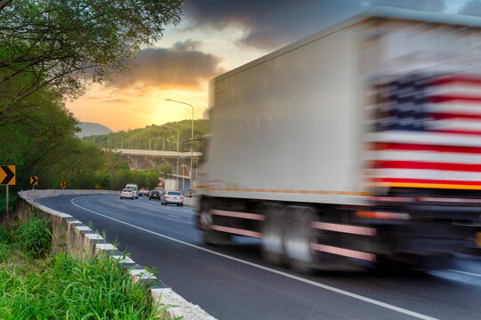 Motion Blur Of Truck With Space For Text And USA Flag On End Cap Driving Fast On The Countryside Road With Mountain Against Sunset