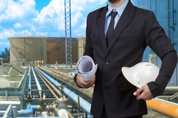 Engineer holding hard hat and blueprint for working at pipe line connection to oil tanks in petrochemical oil refinery