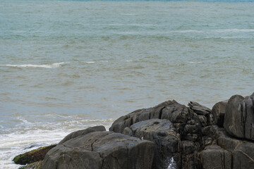 View of the beach of Rio das Ostras in Rio de Janeiro with sunny day, blue sky and some clouds. Strong sea and yellowish sand and lots of rocks.