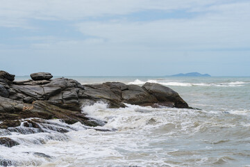 View of the beach of Rio das Ostras in Rio de Janeiro with sunny day, blue sky and some clouds. Strong sea and yellowish sand and lots of rocks.