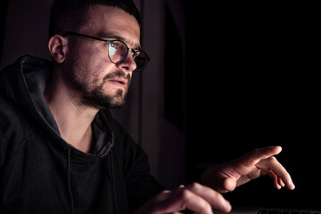 A man with glasses in the dark looks at the computer screen, copy space.