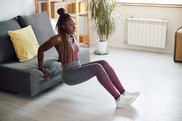African American sportswoman does triceps dips while leaning on sofa at home.