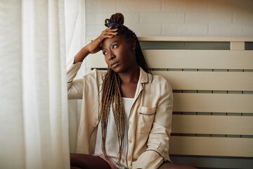 Young sad African American woman looks through the window at home.