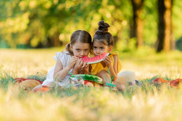 Girls are sitting on a blanket in the park and eat one slice of watermelon. Kids eat fruit outdoor. Healthy snack for children. Little girls are playing in the forest, biting a slice of watermelon.