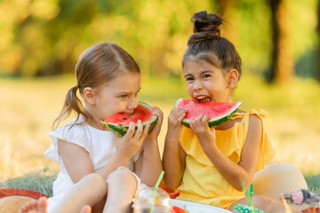 Two little mixed race kids consist of european and caucasian girls smiling with happiness, fun amusement, playing, sitting for picnic and eating piece of watermelon fruit in outdoor garden