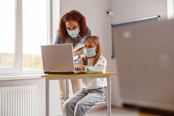 Teacher and schoolgirl in masks using tablet in classroom