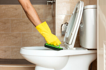 Human hand in a yellow rubber glove is using a plastic brush to scrub the toilet bowl.