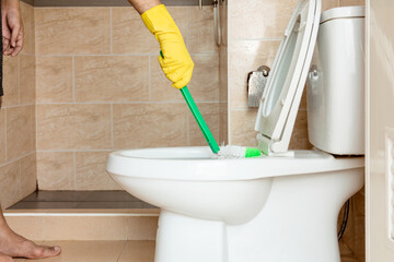 Human hand in a yellow rubber glove is using a plastic brush to scrub the toilet bowl.