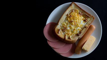 Abstract dark tone of food. Breakfast set includes Egg toast topped with cheese. with chicken ham, sausage and sweet egg. In a white plate and placed on a black tiled background.