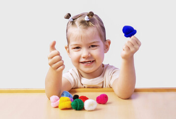 A cheerful girl with doll knitted hats on her hands on a white background. A little girl is sitting at a table and playing games with tiny funny toys on her fingers.  Children's games. Happy childhood