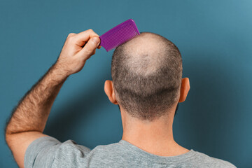 A man combs his bald head with a comb. Blue background. Rear view. The concept of alopecia and...