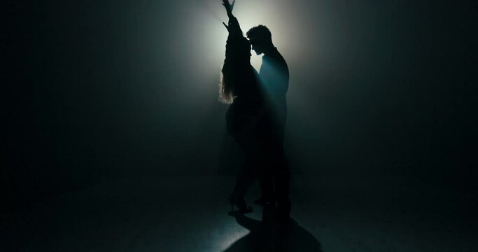 Ballroom dance between two partners shadows of figures on stage in dark room lit by lamp, sexy movements of blonde woman in stilettos, man dressed in black shirt holds partner