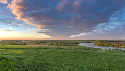 Obraz premium Scenic view of a beautiful summer river sunset with reflection in the water, calm water. Bright colorful cloudy sky. Lush green grass in the foreground.